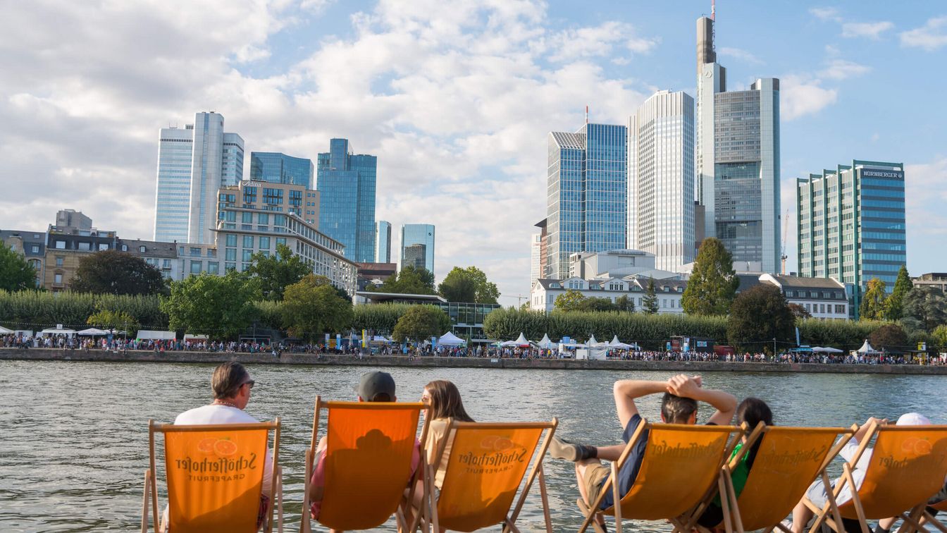 Menschen sitzen in Liegestühlen an Mainufer und blicken auf die Skyline