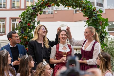 Die Krönung der neuen Frankfurter Weinkönigin unter einem Blumenbogen: Zwei Frauen in Trachten setzen der strahlenden Weinkönigin die Krone auf, während eine feiernde Gruppe zuschaut.