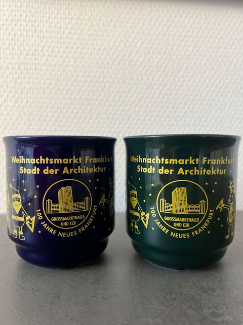 Zu sehen sind zwei Tassen in blau und grün. In der Mitte der Tassen ist in gold ein Kreis und dort zu sehen sind zwei Gebäude, wobei eins von ihnen ein Hochhaus ist. Darunter steht Grossmarkthalle und EZB. Unter dem Kreis list man 100 Jahre Neues Frankfurt. Neben dem Kreis steht links ein Mann mit Bart und einem Winkel in der Hand. Ebenfalls in gold skizziert. Rechts vom Kreis ist ein Rentier mit einem Zirkel in der Hand. Auch in Gold dargestellt. Über dem Kreis steht Weihnachtsmarkt Frankfurt Stadt der Architektur. 