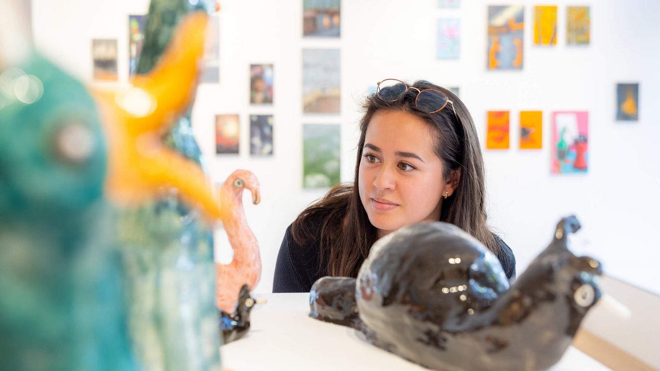 Young woman in a gallery, looking at art, a snail made of clay. In the background on the wall colourful small pictures are hanging.