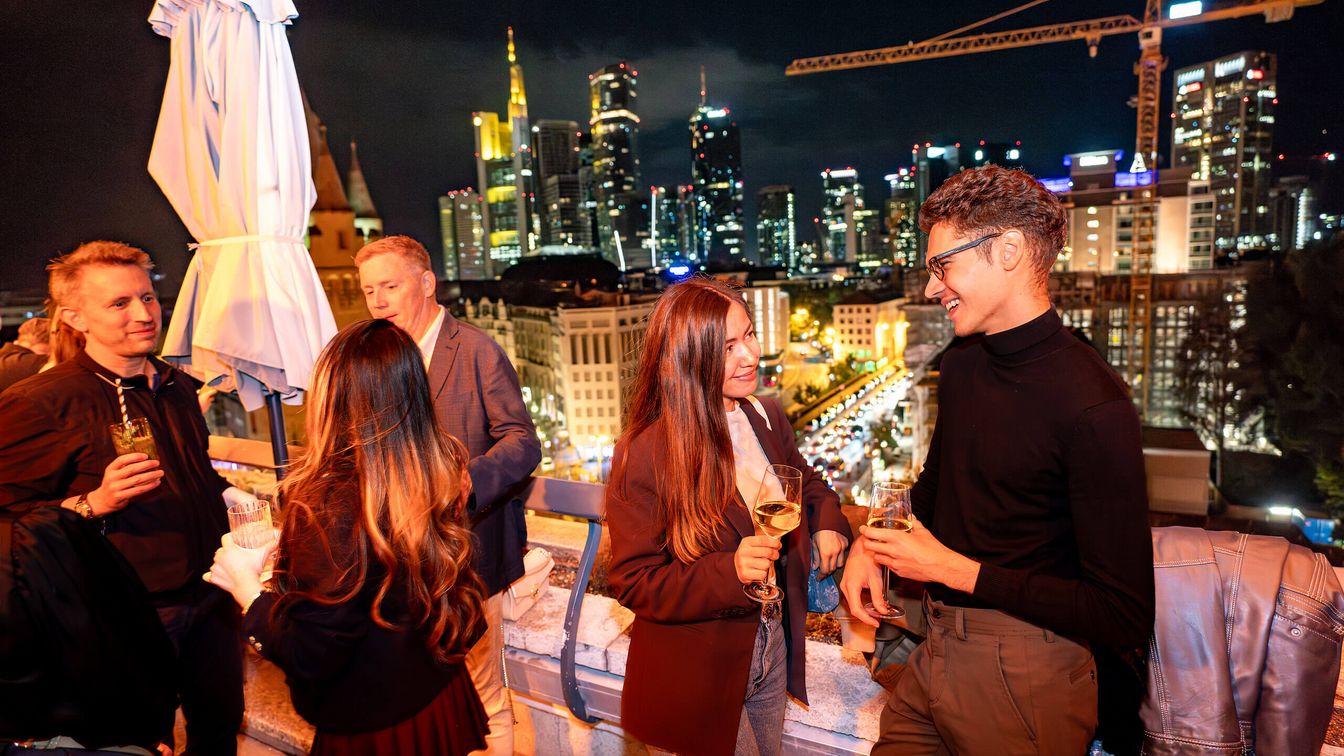 Nachtaufnahme einer Gruppe auf einer Dachterrasse mit Stadtlichtern.