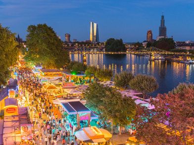 Marktstände und bunt beleuchtete Buden am Mainufer, mit Menschenmassen und Brückenlichtern bei Abendstimmung.