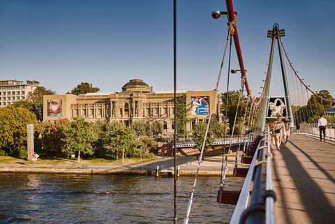 Blick auf das Städel Museum in Frankfurt mit prächtiger Außenfassade, umgeben von Bäumen, im Vordergrund der Holbeinsteg mit Spaziergänger*innen über dem Main.