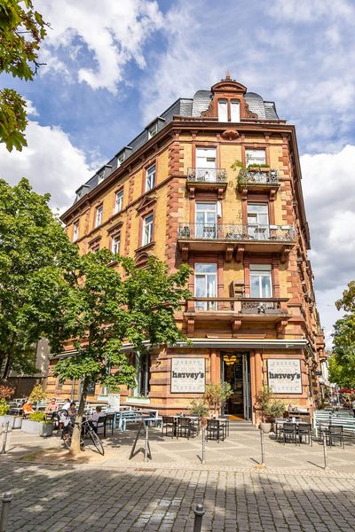 Historisches Gebäude mit Balkonen und Café im Erdgeschoss, umgeben von Bäumen und Straßenflair.