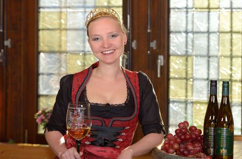 Weinkönigin steht mit Weinglas neben Trauben und Weinflaschen.