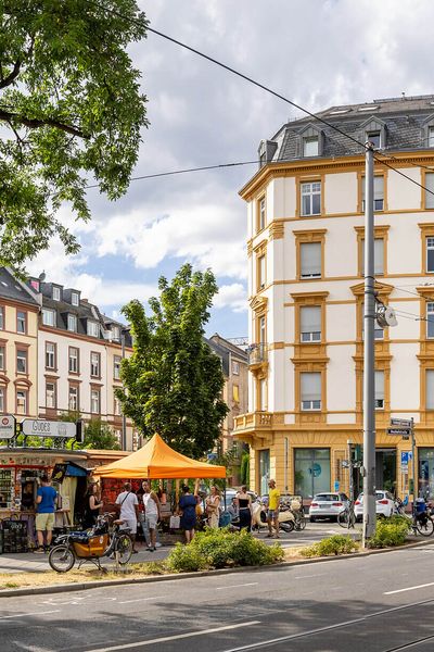 Eine Kreuzung an der Friedberger Landstraße im Frankfurter Nordend, flankiert von historischen Gebäuden und Bäumen. Menschen genießen den Sommer in einem kleinen Verkaufsstandbereich.