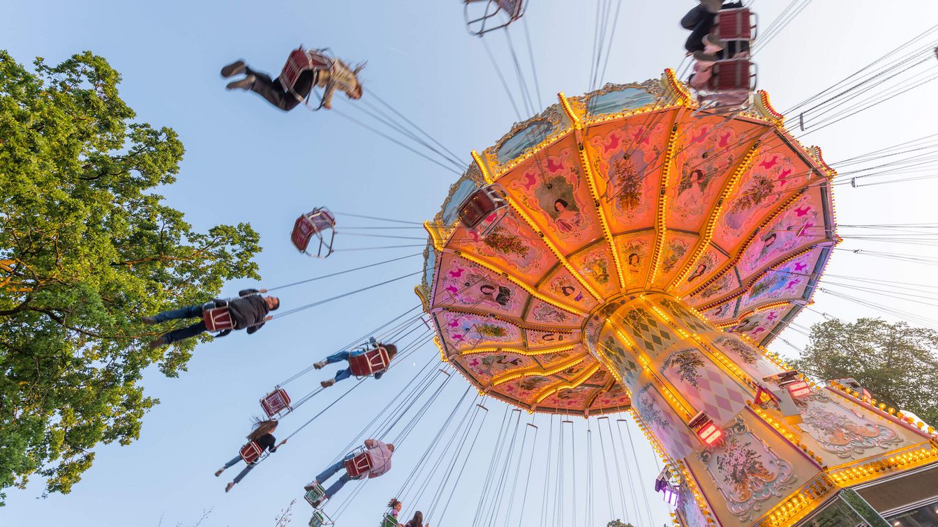 A glowing chain carousel at Frankfurt's Wäldchestag. People float in the air against a blue sky and green trees.
