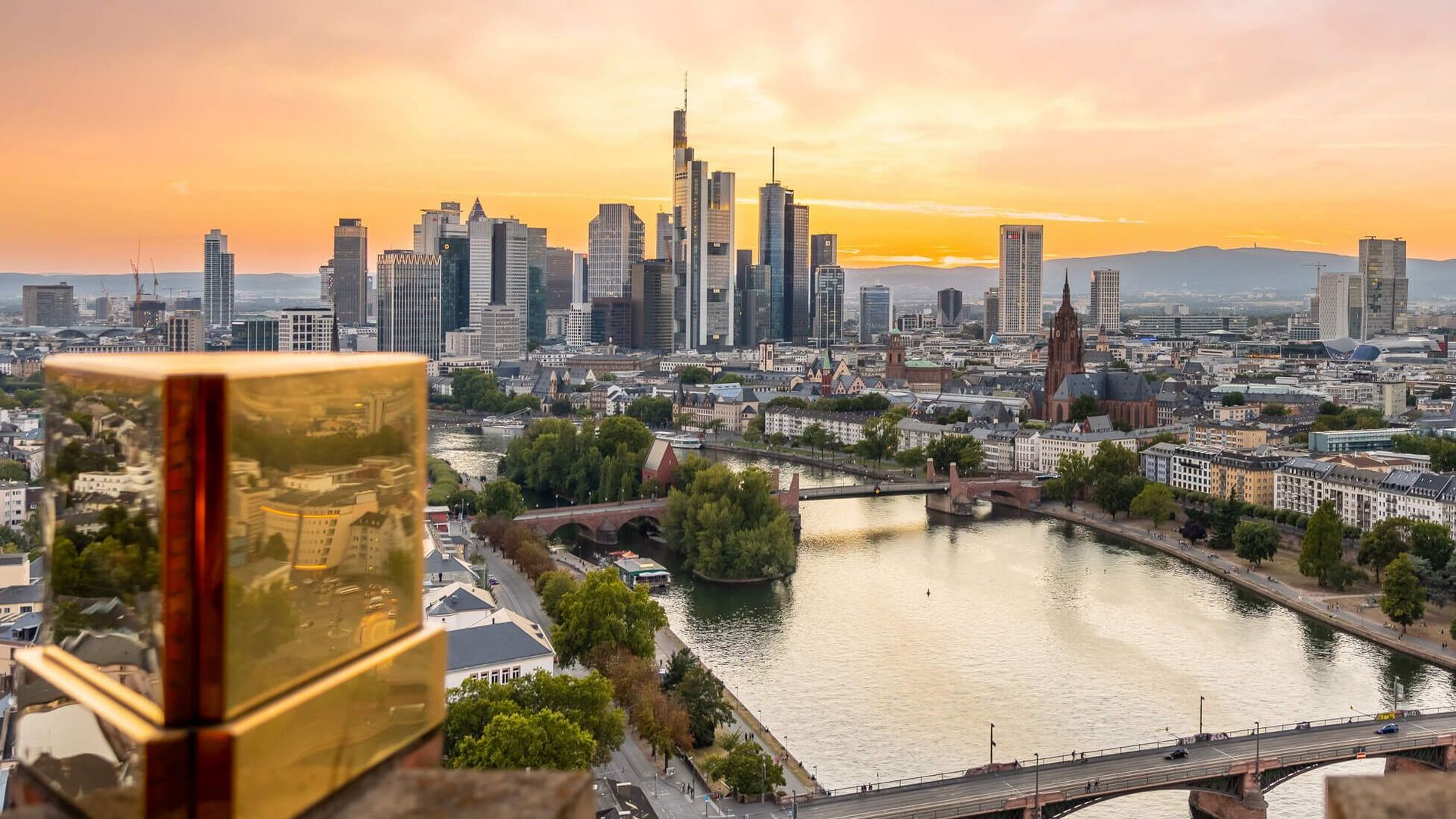 Blick auf den Main, die Frankfurter Skyline und den Kaiserdom vom Dach des Lindner Hotel Frankfurt Main Plaza bei Sonnenuntergang, im Vordergrund goldene Zierelemente des Hoteldachs.