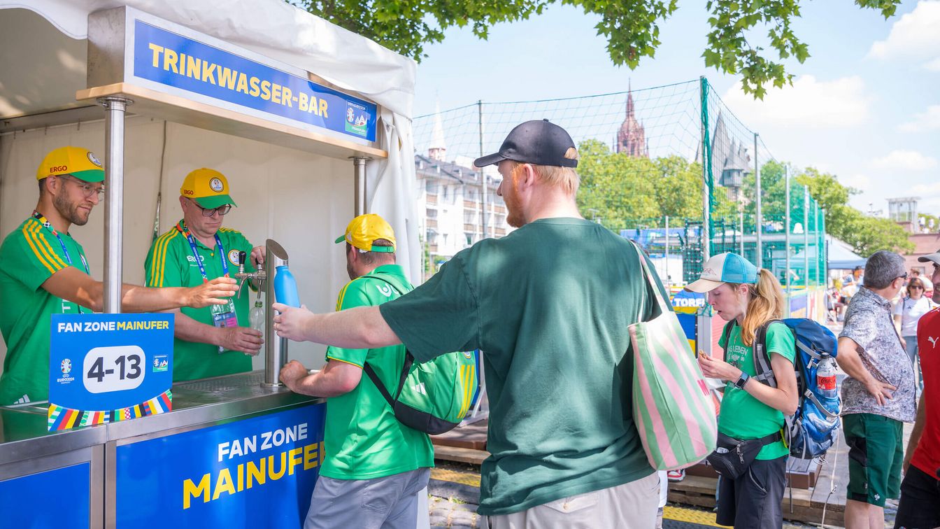 Gäste der Fan Zone und Volunteers stehen an einer Trankwasser-Bar an um sich frisches Wasser geben zu lassen. 
