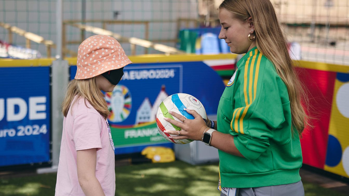 Ein Volunteer erkläre einem Mädchen mit Brille zur Sichteinschränkung wie sie Fußball mit einem Goal Ball spielen kann. 