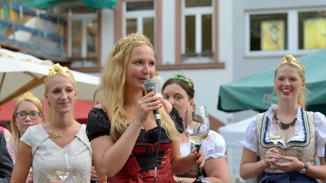 Frau mit Krone spricht ins Mikrofon bei Veranstaltung mit Weinköniginnen.