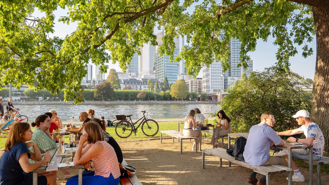 Einige Gäste sitzen im Sommer auf Holzbänken im Maincafé am Ufer des Mains.